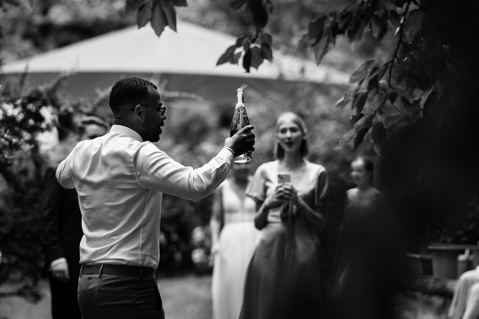 NinaundGeorg 4 Bruder der Braut öffnet eine Sektflasche nach der Trauung im Schlossgarten Starnberg, eingefangen in dynamischer Schwarz-Weiß-Reportage.