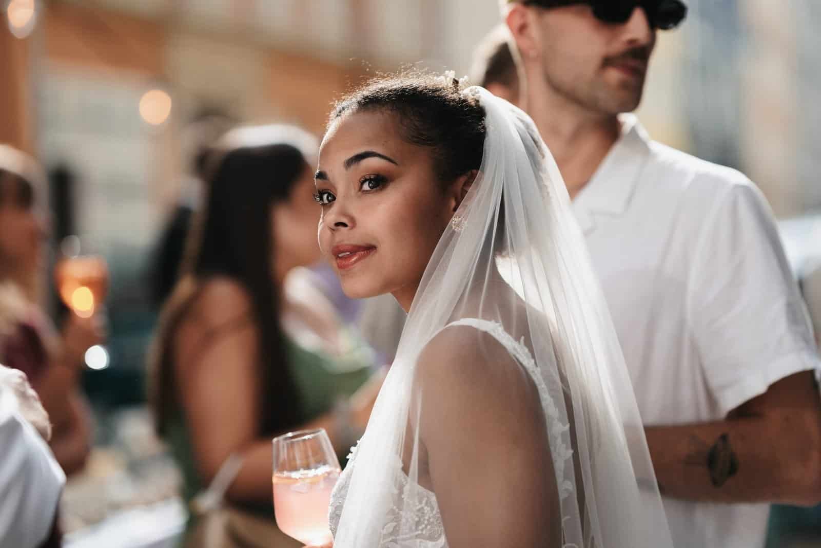 Die Braut lächelt mit einem Drink in der Hand über die Schulter, entspannter Outdoor-Empfang vor der Hochzeitslocation Anju in München.
