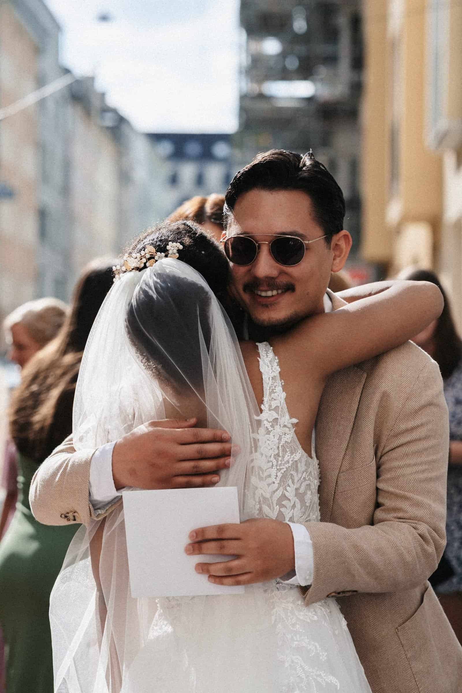 Braut wird von Gast auf der Terrasse der Hochzeitslocation in München freudig umarmt, urbane Reportage.